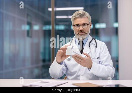 Leitender männlicher Arzt in Uniform hält eine weiße Pille Flasche, während er während eines Videogesprächs spricht, eine Telehealth Verordnung erklärt und professionelle medizinische Fernberatung bietet Stockfoto