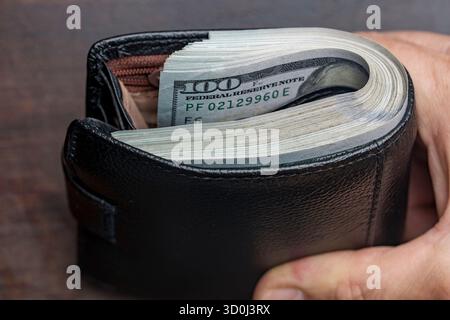 Die Hand eines Mannes hält eine schwarze Ledertasche mit einem Haufen 100-Dollar-Scheine, Banknoten, Bargeld, Nahaufnahme. Konzept für verlorenes oder gefundenes Wallet mit CA Stockfoto