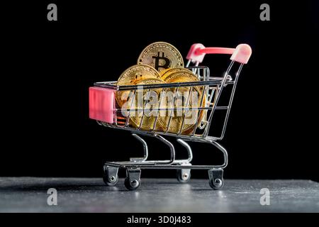 Kryptowährungs-goldene Bitcoin-Münzen in einem kleinen Einkaufswagen auf schwarzem Hintergrund, Nahaufnahme. Digitales Geld und Bitcoin-Konzept Stockfoto