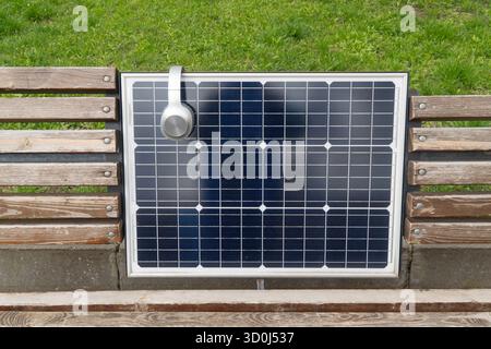 Kopfhörer auf einem dunkelblauen Solarpanel, der in eine Holzbank integriert ist, stehen für erneuerbare Energien und öffentliche Ladetechnik im Freien Stockfoto