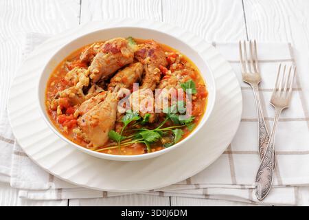 Chakhokhbili, georgianischer Hähncheneintopf mit Tomaten, Kräutern und gehackten Walnüssen in einer Schüssel auf einem weißen Holztisch mit Gabeln, horizontale Ansicht, Nahaufnahme, nicht Stockfoto