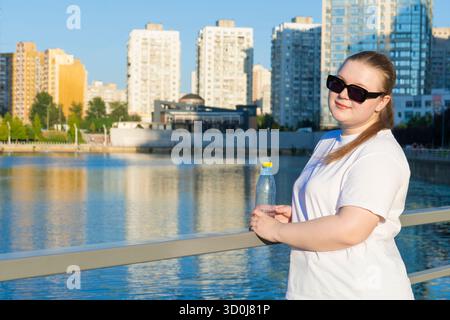 Porträt einer lächelnden jungen Frau in Übergröße in Sonnenbrille, die eine Plastikflasche am Fluss hält, mit einem modernen Stadtbild hinter sich Stockfoto