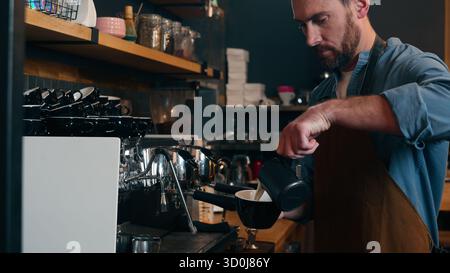 Professioneller kaukasischer Arbeitgeber Barkeeper Mann, der die Bestellung im Café-Shop vorbereitet männlicher Barista-Typ, der Milchcreme in Tasse Latte Cafeteria für Kleinunternehmen gießt Stockfoto
