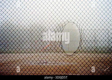Analog poetic shot of an empty concrete mini football field with a wire fence featuring a rounded hole in the foreground, foggy winter morning. Stockfoto