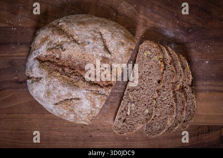 In Scheiben geschnittenes, rundes Sauerteigvollkornbrot mit Walnüssen auf dunklem Holzbrett, Ansicht von oben mit Textur und Nussstücken. Stockfoto