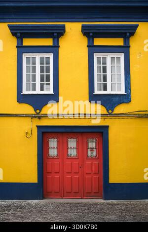 Farbenfrohe Fassade eines Gebäudes mit hellgelben Wänden, blauen Fensterrahmen und einer roten Tür, die lebendigen architektonischen Kontrast und urbanem Charme bietet Stockfoto