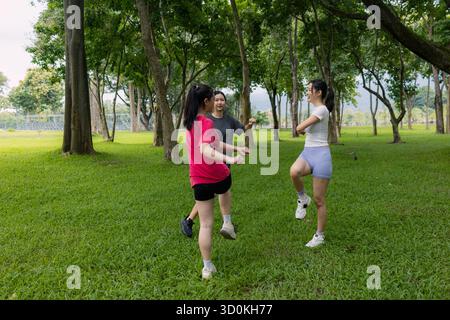 Drei Frauen trainieren gemeinsam in einem üppig grünen Park und genießen ein aktives Outdoor-Workout unter hohen Bäumen. Sie wirken fröhlich und fokussiert und fördern fr Stockfoto