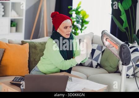 Seniorin, die sich zu Hause durch eine Heizung warm hält, in kuscheliger Winterkleidung gekleidet, bietet Komfort und Entspannung in Innenräumen. Stockfoto