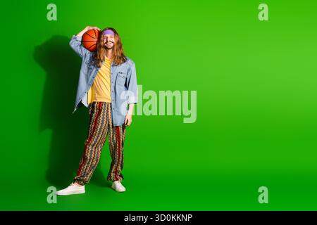 Trendiger junger Mann, der Basketball im stilvollen Outfit auf hellgrünem Hintergrund mit böhmischen Vibes hält Stockfoto