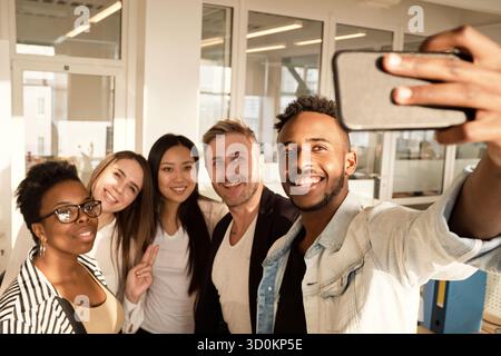 Multiethnische Gruppe junger Kollegen lächelt fröhlich auf die Smartphone-Kamera, während sie im modernen Büro Selfie machen Stockfoto