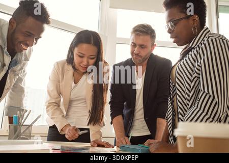 Multiethnisches Team von jungen Managern, die an einem neuen Projekt im Konferenzraum arbeiten, um den Tisch stehen und Brainstorming machen, aus einem niedrigen Winkel sehen Stockfoto