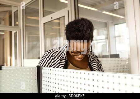 Schwarze Frau in gestreifter Bluse, die in einem modernen Büro an der Trennwand steht, nach unten blickt und lächelt Stockfoto