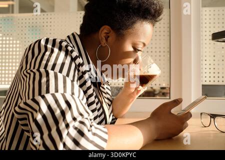Profilansicht einer stilvollen afroamerikanischen Frau in schwarz-weiß gestreifter Bluse, die am Bürotisch sitzt, Kaffee trinkt und das Smartphone benutzt Stockfoto