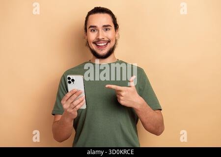 Glücklicher junger Mann mit Smartphone, das auf den Bildschirm zeigt und ein grünes T-Shirt vor beigefarbenem Hintergrund für Freizeit-Lifestyle-Shopping und Werbung trägt Stockfoto