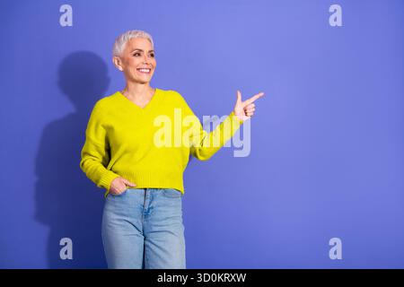 Eine ältere Frau in einem hellgelben Pullover lächelt, als sie vor einem lila Hintergrund nach rechts zeigt Stockfoto