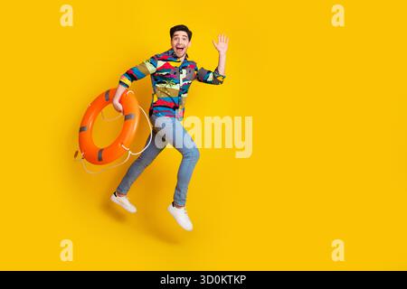 Junger Mann, der mit Rettungsschirm auf hellgelbem Hintergrund springt, der buntes Hemd für Lifestyle-Werbung und Modeschaffung trägt Stockfoto