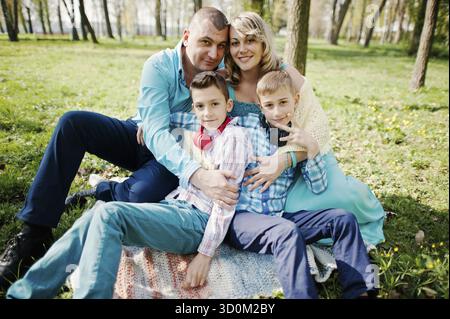 Glücklich schwanger Familie mit zwei Söhnen, gekleidet in eine türkise Kleidung, sitzen auf dem Rasen mit Blumen im park Stockfoto