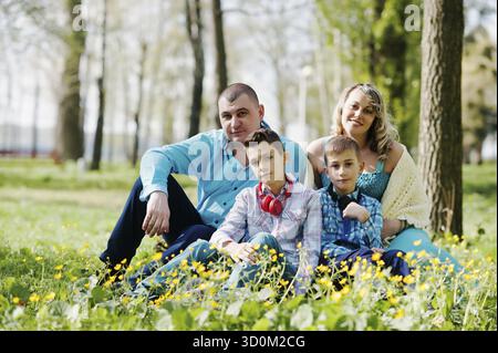Glücklich schwanger Familie mit zwei Söhnen, gekleidet in eine türkise Kleidung, sitzen auf dem Rasen mit Blumen im park Stockfoto