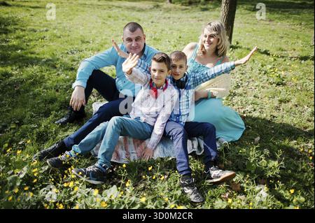 Glücklich schwanger Familie mit zwei Söhnen, gekleidet in eine türkise Kleidung, sitzen auf dem Rasen mit Blumen im park Stockfoto