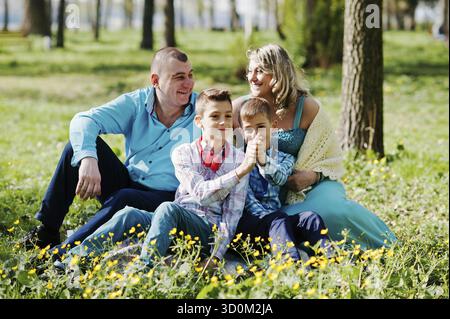 Glücklich schwanger Familie mit zwei Söhnen, gekleidet in eine türkise Kleidung, sitzen auf dem Rasen mit Blumen im park Stockfoto