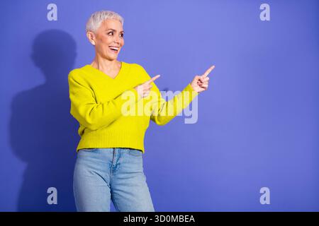 Reife Frau mit kurzem grauem Haar Wear Pullover zeigt vor einem lila Hintergrund, lächelt warm und fesselt den Zuschauer in einen lässigen Lebensstil Stockfoto