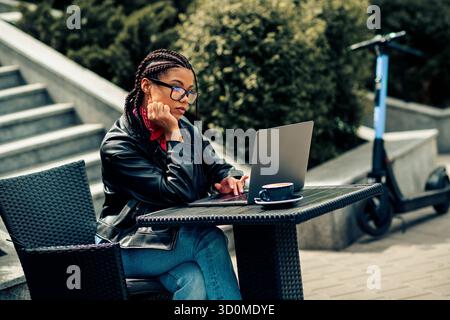 Eine junge Frau mit geflochtenen Haaren nutzt ihren Laptop im Freien und genießt die städtische Umgebung und die entspannte Freizeit am Frühlingstag Stockfoto