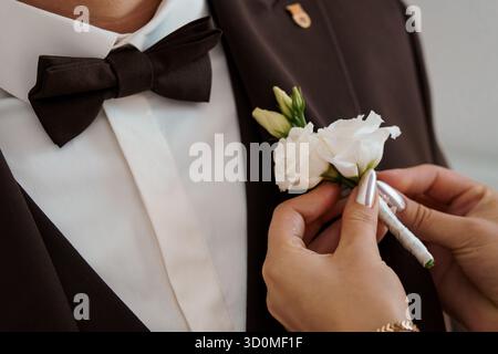 Eine Nahaufnahme zeigt eine Person, die akribisch eine zarte weiße Blumenkuppe an das Revers eines dunkelbraunen Smoking des Bräutigams heftet und ein R zeigt Stockfoto
