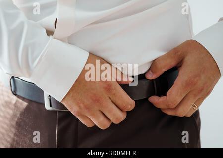 Ein Nahbild zeigt einen Mann, der einen dunkelbraunen Ledergürtel mit einer silbernen Schnalle um die Taille über einer grauen Hose hält, was auf ein formelles OR hindeutet Stockfoto
