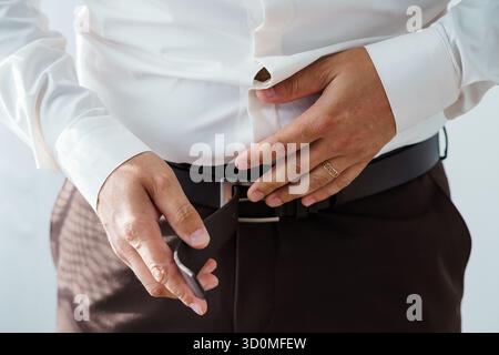 Eine Nahaufnahme zeigt einen Mann, der einen schwarzen Ledergürtel anlegt, ein weißes Hemd und eine karierte Hose trägt und einen silbernen Ring und einen professionellen dre zeigt Stockfoto