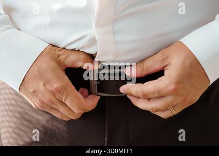Eine Nahaufnahme zeigt einen Bräutigam, der seinen braunen Ledergürtel während der Hochzeitsvorbereitungen mit einer schwarzen Schnalle sichert. Konzentrieren Sie sich auf Hände, formelle Kleidung und inti Stockfoto
