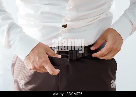 Eine Nahaufnahme zeigt einen Mann, der einen dunklen Ledergürtel um seine Taille hält, braune Hosen und ein helles Hemd trägt, was auf formelle oder bu hindeutet Stockfoto