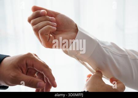Eine Nahaufnahme zeigt einen Mann, der Hilfe beim Befestigen silberner Manschettenknöpfe an seinem knusprigen weißen Hemd erhält, was auf formelle Geschäftskleidung und hinweist Stockfoto