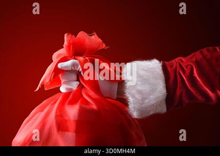 Die Hand des Weihnachtsmanns in einem Anzug und Handschuhen hält einen roten Sack mit Geschenken, die zu Weihnachten verteilt werden. Stockfoto