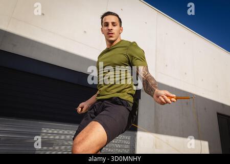 Ein junger, sportlicher Mann, der vor einer Betonwand ein Sprungseiltraining macht. Fotografiert im Sommer unter warmem Licht. Stockfoto