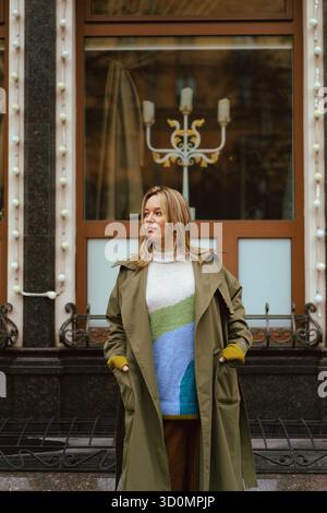 Modische Frau im Trenchcoat, die vor einem verzierten Fenster steht Stockfoto