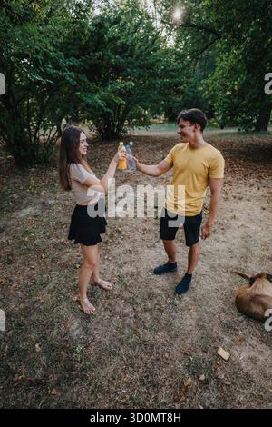 Junges Paar, das in einem sonnigen Park mit Drinks toast, im Freien, zwangloser Dates-Moment Stockfoto