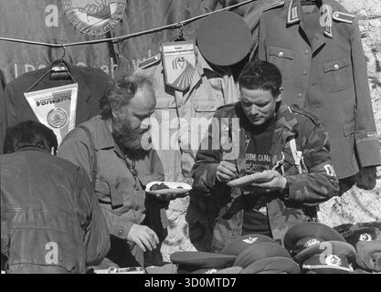 DEU, Deutschland, Berlin, 18.03.1990: Am ehemaligen Checkpoint Charlie machen Händler, die DDR-Erinnerungsstücke aller Art an Touristen verkaufen, eine Pause mit einer Currywurst. Stall mit Uniformen, Wimpeln, Militärabzeichen und Uniformmützen. Ein Käufer am Stand. [Automatisierte Übersetzung] Stockfoto