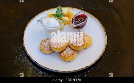 Hüttenkäsepfannkuchen, Curdish Pancakes, Käsekuchen, Syrniki mit frischer Marmelade, Sauerrahm zum Frühstück. Käsepfannkuchen mit Kirschmarmelade, Sauerrahm und Minze auf einem Teller. Hohe Qualität Stockfoto
