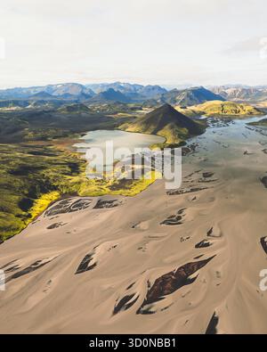 Aus der Vogelperspektive ein starker Kontrast zwischen dem dunklen, sandigen Flussbett und den lebhaften grünen Hügeln, die einen ruhigen See umgeben, F208, Island. Stockfoto