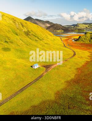 Blick aus der Vogelperspektive auf ein einsames weißes Haus, eingebettet in lebendige grüne Hügel, die rostfarbene Ebenen unter einem riesigen blauen Himmel treffen, F208, Island. Stockfoto
