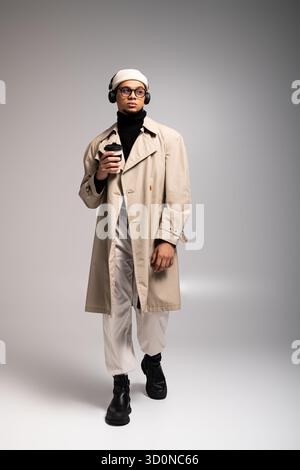 Ein junger Mann läuft selbstbewusst in einem modischen Outfit, schlürft Kaffee und trägt Kopfhörer. Stockfoto