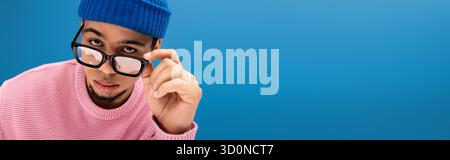 Ein eleganter junger Mann passt seine Sonnenbrille an und trägt einen trendigen Pullover vor blauem Hintergrund Stockfoto