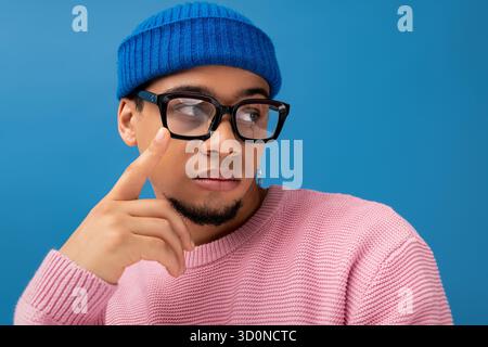 Ein junger Mann in einem pinkfarbenen Pullover und einer blauen Beanie posiert nachdenklich und unterstreicht seinen modischen Look. Stockfoto