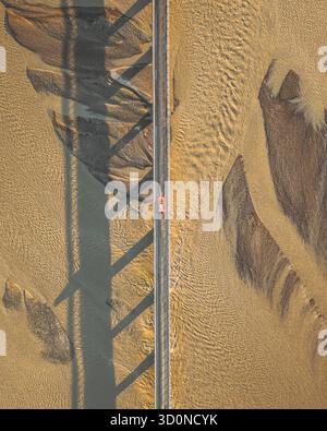 Aus der Vogelperspektive eines einsamen roten Autos, das eine lange Brücke über strukturiertem, sandigem Gelände unter starken Schatten überquert, Stafafafell, Sveitarfélagið Hornafjörður, Island. Stockfoto