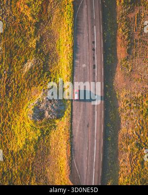 Aus der Vogelperspektive eines einsamen roten Autos, der eine schroffe Straße überquert, flankiert von Feldern aus Gold und Grün, und durch die einsame und dennoch wunderschöne Landschaft in Grundarfjörður, Island. Stockfoto
