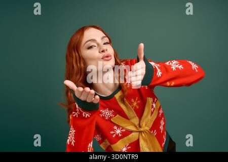 Eine rothaarige Frau posiert spielerisch in einem festlichen Weihnachtspullover gegen Grün. Stockfoto