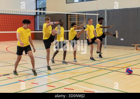 Verschiedene männliche Volleyball-Teamkollegen, die hohe Beine im Fitnessstudio mit Volleyballnetz und Ball spielen. Athleten, Teamwork, Fitness, Sport, Wettkampf, Dynamic, activewear || Modell veröffentlicht Stockfoto