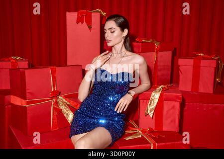 Schöne junge Frau sitzt anmutig in festlicher Kleidung, umgeben von leuchtenden roten Geschenkboxen. Stockfoto
