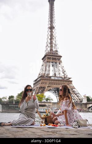 Zwei Frauen, die sich beim Picknick vor dem Eiffelturm mit dem Smartphone entspannen || Model veröffentlicht Stockfoto