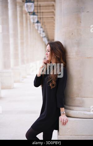 Stilvolle junge Frau in schwarzem Kleid, die sich nachdenklich auf eine Steinsäule im Palais Royal in Paris, Frankreich, im Freien stützt. || Modell freigegeben Stockfoto